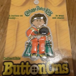 Puede incluir: Un botón coleccionable de Cabbage Patch Kids con un personaje de dibujos animados que lleva un chándal rojo y verde y sostiene una raqueta de tenis. El botón está etiquetado como "Buttonons" e incluye un botón para una camisa.