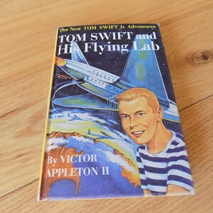 Pode incluir: Um livro vintage intitulado "TOM SWIFT and His Flying Lab" de Victor Appleton II. A capa apresenta um jovem sorrindo com uma camisa listrada e um avião futurista contra um céu estrelado. O livro tem uma lombada amarela.