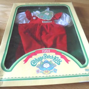 May include: A 1984 Cabbage Patch Kids doll outfit in original packaging, featuring a red overall-style garment with white puffed sleeves. The box displays the text Cabbage Patch Kids, 1984, and Fits All 16 inch Kids, Ages 3 & Up, Coleco.