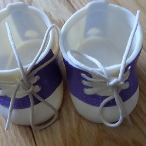 Puede incluir: Un par de zapatos de muñeca blancos y morados con cordones blancos. Los zapatos son de plástico y tienen un diseño sencillo.