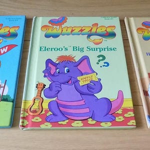 Op de afbeelding: Drie kinderboeken met kleurrijke illustraties. De boeken heten "Wuzzles" en bevatten stripfiguren. De titels zijn onder meer "Win One For Bumblelion", "Eleroo's Big Surprise" en "Hoppopotamus Plays Detective". De boeken liggen op een houten oppervlak.