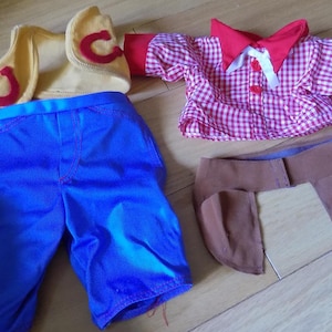 May include: A set of doll clothing including a yellow waistcoat with red horseshoe appliqués, blue trousers, a red and white gingham shirt with a red collar and cuffs, and a brown fabric accessory piece with two hanging panels and a central strap.