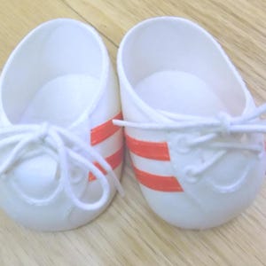 Puede incluir: Un par de zapatos de bebé blancos con rayas naranjas y cordones blancos. Los zapatos están hechos de un material suave y están diseñados para parecerse a zapatillas deportivas. Los zapatos están sobre una superficie de madera.