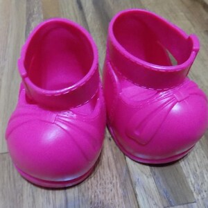 Puede incluir: Un par de zapatos de muñeca de plástico rosa brillante. Los zapatos tienen un estilo Mary Jane con una correa en la parte superior y un detalle de lazo. Los zapatos están sobre una superficie de madera.