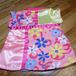 May include: A doll dress featuring a pink floral pattern with blue, yellow, and white flowers. The dress has pink satin panels and trim, with a yellow satin sleeve and a pink collar.