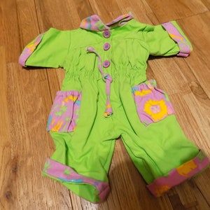 Peut inclure: Une grenouillère bébé verte avec des bordures roses et jaunes et des motifs d'animaux de dessin animé. La grenouillère a un cordon de serrage à la taille et une fermeture à bouton au cou.