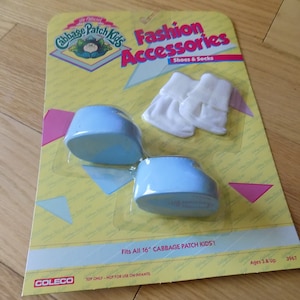 Może przedstawiać: Opakowanie akcesoriów modowych Cabbage Patch Kids dla lalek o wysokości 40,6 cm. Opakowanie zawiera parę niebieskich butów i parę białych skarpet. Opakowanie jest żółte z różowymi akcentami i zawiera logo Cabbage Patch Kids.