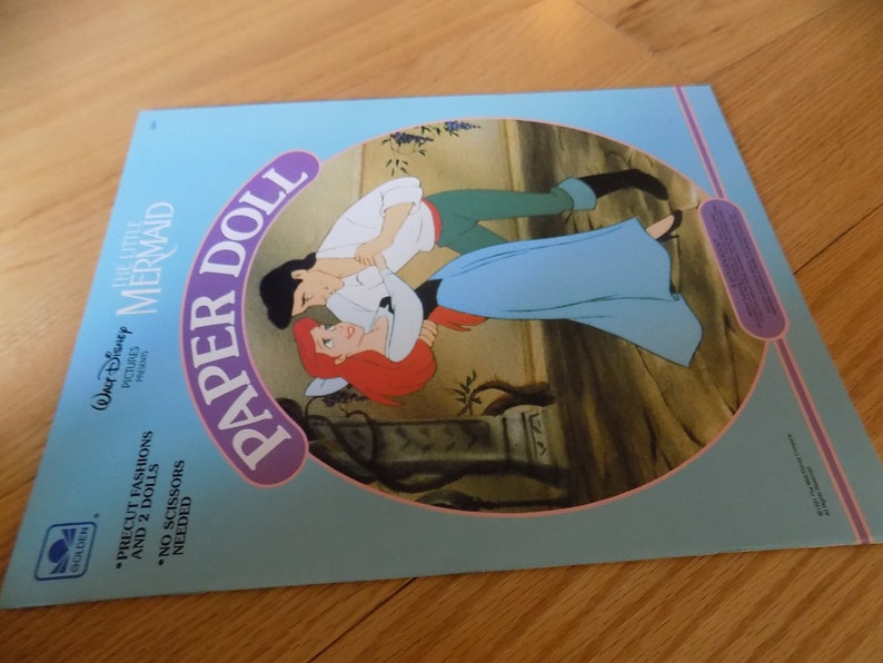 May include: A blue and pink paper doll book featuring Ariel and Eric from Disney's The Little Mermaid. The cover shows Ariel and Eric dancing together. The book includes precut fashions and two dolls. No scissors are needed.