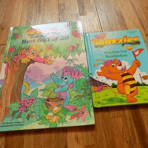 Op de afbeelding: Twee kinderboeken. Het eerste boek is getiteld "and the Best Gift of All" en toont een cartoon-aap en een blauwe draak. Het tweede boek is getiteld "Wuzzles" en toont een cartoon-leeuw met een bijgestreept lichaam. De tekst op het tweede boek luidt "Win One For Bumblelion".
