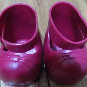 Pode incluir: Um par de sapatos de boneca de plástico vermelho com um detalhe de laço. Os sapatos têm uma tira de tornozelo.