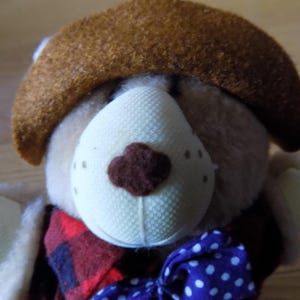 Puede incluir: Un oso de peluche con un sombrero marrón, una nariz blanca y una bufanda a cuadros rojos y negros. El oso tiene una pajarita azul con lunares blancos. El pelaje del oso es de color marrón claro.