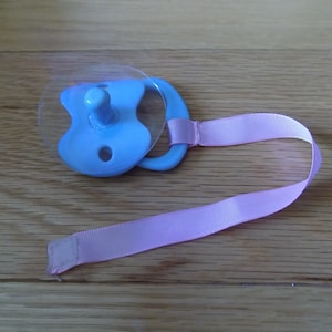 Pode incluir: Uma chupeta azul com um escudo de plástico transparente e uma fita rosa amarrada.