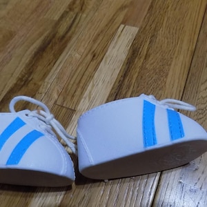 Puede incluir: Un par de zapatillas blancas con rayas azules. Las zapatillas están hechas de un material suave y blanco y tienen un diseño simple.