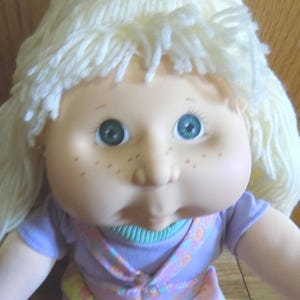 Puede incluir: Una muñeca Cabbage Patch Kid con pelo de lana rubio, ojos verdes y un vestido floral morado y rosa. La muñeca tiene una tez rosada y pecas.