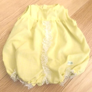 May include: A yellow baby romper with a gingham pattern. The sleeveless romper has white lace trim down the centre and around the leg openings. The fabric is a light yellow with a small white check pattern. A small tag is visible.