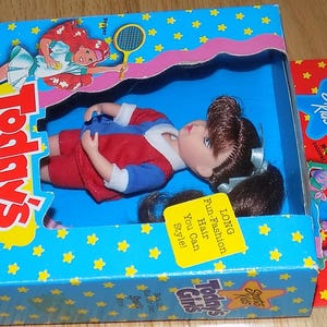May include: A doll with long brown hair wearing a red and blue sports uniform. The doll is packaged in a blue and yellow box with the words "Today's Girls Sports Kids" on it. The box also includes a smaller card with the words "Today's Girls Sports Kids" on it.