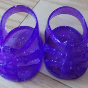 Puede incluir: Un par de sandalias de gelatina de color morado translúcido con un acabado brillante, diseño de punta redondeada y una sola correa en el tobillo. Estos zapatos pequeños de plástico tienen una parte superior tipo jaula con varios recortes.
