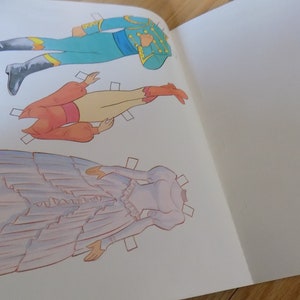 May include: A paper doll set with three figures. The figures are a man in a blue and yellow uniform, a woman in a red and white dress, and a woman in a purple dress. The figures are all cut out and ready to be played with.