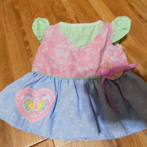 May include: A light blue and pink baby dress with a heart-shaped patch featuring two fish and the text "First Kiss CPK". The dress has a green and white polka dot bodice and a light blue skirt with a white pattern.