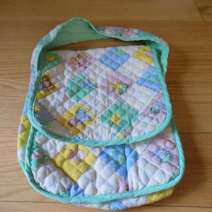 Puede incluir: Bolso de hombro acolchado con diseño patchwork en colores pastel. El bolso presenta un cierre de solapa y un ribete y correa verde menta. El patchwork incluye cuadrados con ilustraciones de temática infantil.