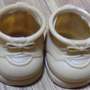 Puede incluir: Un par de zapatos de muñeca de color marrón claro con un lazo en la parte delantera. Los zapatos tienen un diseño cosido y una punta redonda.