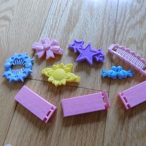 May include: A collection of colorful plastic hair clips, including a blue comb clip, a pink bow clip, a yellow sunflower clip, a purple star clip, a pink comb clip, a blue bird clip, and two pink rectangular clips.