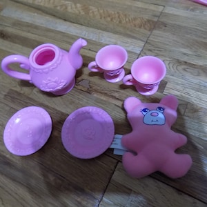 Puede incluir: Juego de té de plástico rosa con una tetera, dos tazas, dos platillos y un oso de peluche rosa.