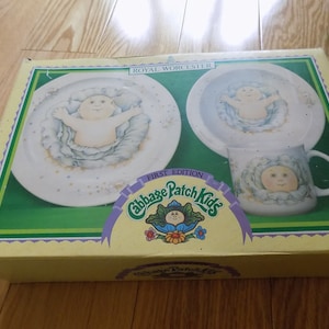 Puede incluir: Una caja amarilla con un borde verde y un logotipo blanco de Cabbage Patch Kids. La caja contiene un juego de tres platos de cerámica blanca con un diseño de Cabbage Patch Kid. Los platos incluyen un plato grande, un plato más pequeño y una taza.