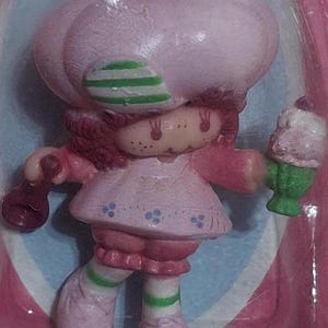 Peut inclure: Une figurine en plastique rose et verte d'une fille avec un chapeau rose et un cache-œil rayé vert et blanc. Elle tient un cornet de glace vert et blanc dans une main et un téléphone rose et blanc dans l'autre.