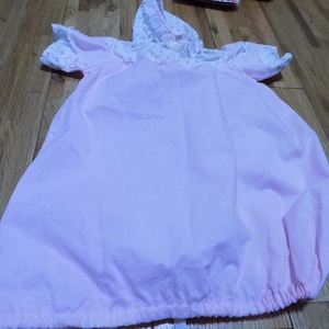 May include: A light pink baby gown with a matching hood. The hood and sleeves are trimmed with white lace and a delicate floral pattern. The gown has a gathered bottom and short sleeves.