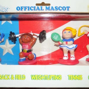 May include: A set of four Olympic themed figurines, each representing a different sport: track and field, weightlifting, tennis, and gymnastics. The figurines are all wearing red, white and blue outfits and have the word "USA" on their uniforms.