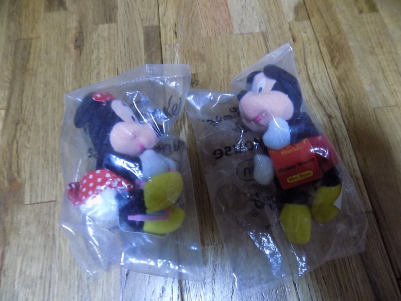 May include: Two plush toys, Mickey Mouse and Minnie Mouse, are packaged in clear plastic bags. Mickey Mouse is wearing red shorts and yellow shoes. Minnie Mouse is wearing a red and white polka dot dress and yellow shoes. The packaging includes the text "World of Disney" and "Mickey Mouse Mini Bean".