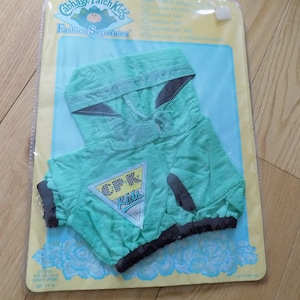 Puede incluir: Una sudadera con capucha verde menta de Cabbage Patch Kids Fashion Separates para muñecas, con ribete gris y un logotipo triangular que dice "CPK Kids". La sudadera está empaquetada en una bolsa de plástico transparente sobre un respaldo de cartón amarillo y azul.