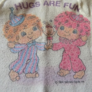 May include: A cream-colored fabric featuring a graphic print of two cartoon characters with curly hair wearing pajamas. The character on the left has orange hair and blue striped pajamas, while the one on the right has pink hair and pink pajamas. The text HUGS ARE FUN is printed above.