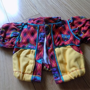Puede incluir: Una colorida chaqueta de muñeca con un patrón de rombos rojos, bolsillos amarillos y ribetes azules. La chaqueta tiene mangas cortas y abullonadas y está abierta por delante. El diseño incluye varios elementos gráficos.