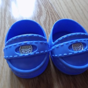 Puede incluir: Un par de zapatos de muñeca de plástico azul con una hebilla plateada que dice "CPK" en cada zapato.