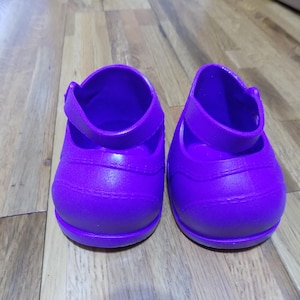 Puede incluir: Un par de zapatos morados brillantes con una correa. Los zapatos están hechos de un material liso y están sobre un suelo de madera. Zapatos para niños.