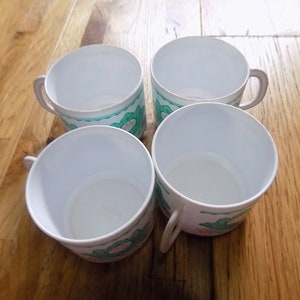 May include: Four white ceramic mugs with green and pink floral designs. The mugs have a rounded shape and a handle on the side.