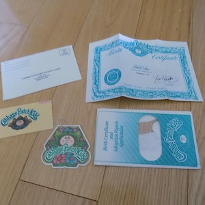 May include: A collection of Cabbage Patch Kids ephemera, including a birth certificate, adoption papers, a tag, and an envelope. The items are on a wooden surface. The birth certificate has a blue border.