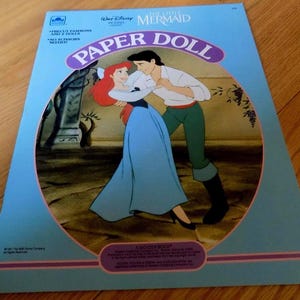 May include: A vintage paper doll book featuring Ariel and Prince Eric from Disney's The Little Mermaid. The cover shows the characters dancing together in a blue and green dress and green pants. The book is titled "The Little Mermaid Paper Doll" and features the text "Precious Fashions for 2 Dolls" and "No Scissors Needed".
