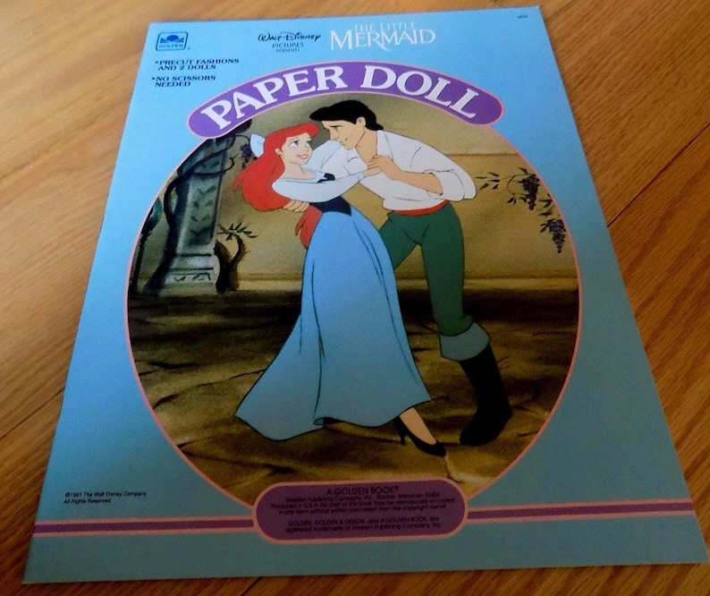 May include: A vintage paper doll book featuring Ariel and Prince Eric from Disney's The Little Mermaid. The cover shows the characters dancing together in a blue and green dress and green pants. The book is titled "The Little Mermaid Paper Doll" and features the text "Precious Fashions for 2 Dolls" and "No Scissors Needed".