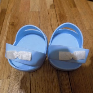 Puede incluir: Un par de sandalias para bebé de color azul claro con lazos blancos. Las sandalias están hechas de un material suave y flexible y tienen un diseño sencillo.