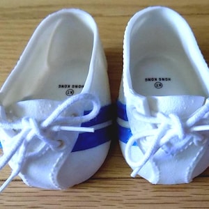 Puede incluir: Un par de zapatos de muñeca blancos con rayas laterales azules y cordones blancos. El interior de cada zapato presenta un logotipo circular con el texto KT y HONG KONG impreso en negro. Los zapatos tienen un diseño de punta redondeada.