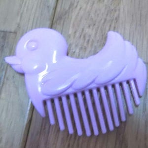 Może przedstawiać: Fioletowy plastikowy grzebień do włosów w kształcie kaczki z rzędem zębów.