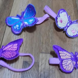 May include: Four embroidered butterfly hair accessories in shades of blue, purple, and yellow. Each butterfly is attached to a matching elastic band or clip. The butterflies have detailed wing patterns and are set against a wooden background.