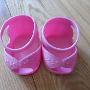 Puede incluir: Un par de zapatos de muñeca de plástico rosa con una correa y un diseño decorativo.