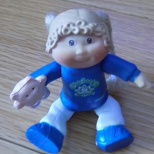 Op de afbeelding: Een Cabbage Patch Kids pop met blond haar in staartjes, die een blauw shirt met een groen logo en een witte broek draagt. De pop houdt een roze varken in haar rechterhand.