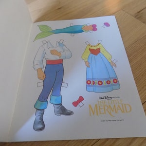 May include: A paper doll set featuring Ariel and Eric from Disney's The Little Mermaid. The set includes Ariel's human form, Eric, and Ariel's tail. The paper dolls are printed on white paper with colorful illustrations.