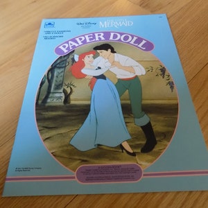 May include: A blue and pink paper doll book featuring Ariel and Prince Eric from Disney's The Little Mermaid. The cover shows Ariel and Eric dancing together. The book includes the text "Paper Doll" in a purple circle.