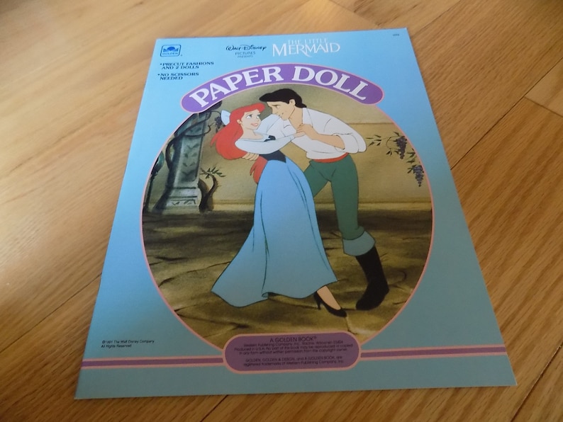 May include: A blue and pink paper doll book featuring Ariel and Prince Eric from Disney's The Little Mermaid. The cover shows Ariel and Eric dancing together. The book includes the text "Paper Doll" in a purple circle.